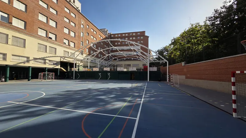 Estructura de arcos de acero sobre la pista deportiva de un colegio, entre edificios de ladrillo