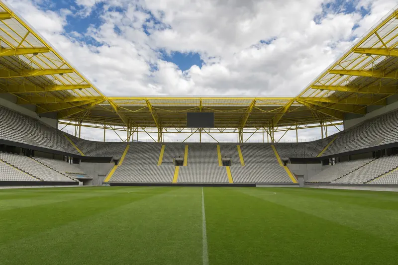 Interior de un estadio vacío con gradas y una gran cubierta de estructura metálica pintada en amarillo (recreación generada con IA)