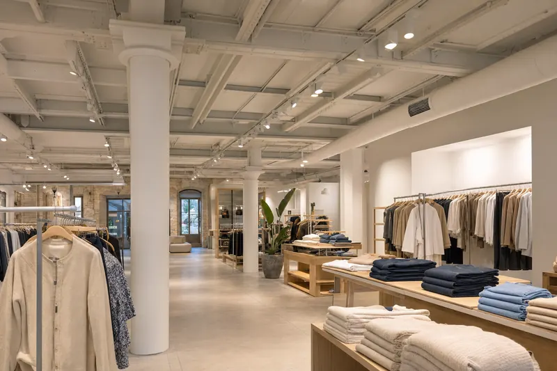 Planta baja de una tienda de ropa con vigas de acero vistas en el techo, conductos de ventilación y focos (recreación generada con IA)